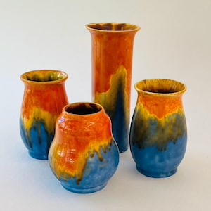 May include: A collection of four ceramic vases featuring a vibrant glaze with blended layers of bright orange, golden yellow, and deep blue. The glaze exhibits a distinct drip pattern where the colours overlap, creating a smooth, glossy finish.