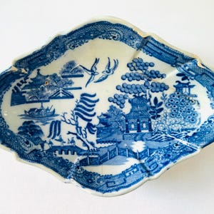 May include: An antique blue and white porcelain dish with an irregular shape. The Willow pattern design includes pagodas, bridges, and birds. The rim has a detailed, patterned border. A vintage decorative piece, ideal for display.