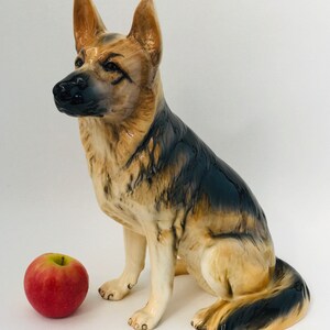May include: A ceramic German Shepherd dog figurine in a seated position, with a red apple beside it. The dog has a black and tan coat, with detailed facial features and a glossy finish. The figurine is a decorative item.