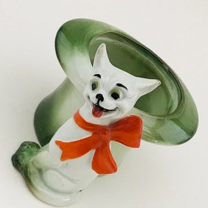 May include: A ceramic figurine of a white dog with a red bow tie, emerging from a green hat. The dog has black eyes and a pink tongue. The hat is a light green color.