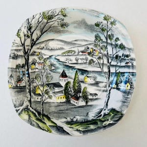 May include: A round decorative plate with a landscape scene of a river, trees, and houses. The plate has a white background with a painted design in shades of grey, blue, yellow, and green. The plate's diameter is approximately 15 cm.
