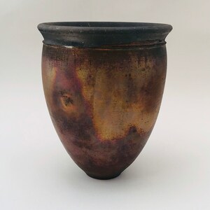 May include: A handmade ceramic vase with a dark brown rim and a pearlescent, metallic finish. The vase has a rounded body and a slightly flared opening. The colours transition from deep burgundy to gold and bronze.