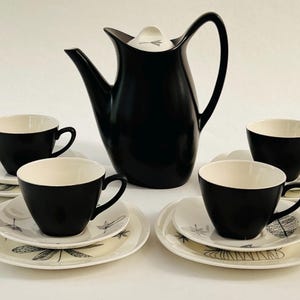 May include: A black and white mid-century modern tea set. The set includes a tall black coffee pot, four black cups with white interiors, and matching saucers with a leaf design. The set is displayed on a white surface.