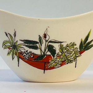 May include: Cream-coloured ceramic bowl with a unique shape. The bowl features a red, boat-shaped design with a botanical illustration of various green plants and white flowers. The bowl's design is outlined in black.