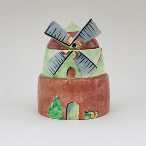 May include: A ceramic windmill figurine with a brown base, green midsection, and brown top. The windmill blades are gray and black. The figurine has a small green tree and a curved doorway.