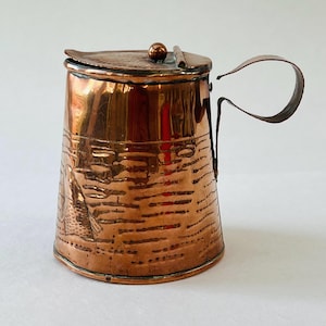May include: A copper pitcher with a lid and handle. The pitcher has a cylindrical body with etched horizontal lines and a fish design. The lid is hinged and has a small ball on top. The handle is a simple loop.