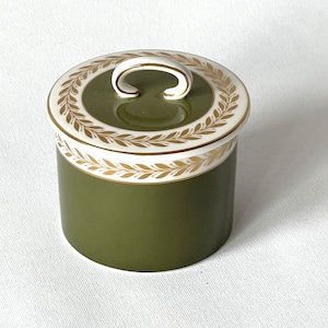 May include: An olive green ceramic jar with a lid. The lid has a white border with gold leaf detailing and a white handle. The jar is cylindrical and sits on a white surface. The jar is likely for storage.