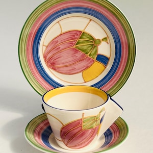 May include: A three-piece ceramic dish set comprising a plate, saucer, and teacup. The set has a cream base with a pink, green, and yellow floral design. The rims are decorated with green, pink, and blue stripes.