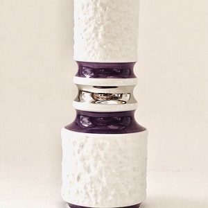 May include: A white ceramic vase with a textured surface and a cylindrical shape. The vase features a purple band at the base and top, with a silver band in the middle. The vase is approximately 10 inches tall.