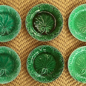 May include: Six green ceramic plates with a leaf design. The plates are arranged in two rows of three, each featuring a detailed embossed leaf pattern in a vibrant green colour. The plates are on a woven, tan surface.