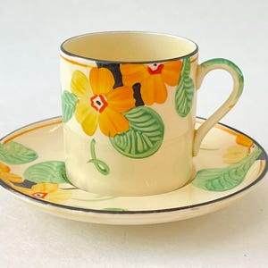 May include: A cream-colored teacup and saucer set. The cup and saucer are decorated with orange and yellow floral designs with green leaves. The rims are outlined in black. The handle of the cup is green.