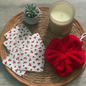 Peut inclure: Un ensemble de quatre disques démaquillants en coton blanc avec un imprimé de cerises rouges, une éponge de bain rouge en forme de cœur, une petite plante succulente en pot et une bougie en verre transparent dans un plateau en osier brun.