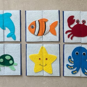 Peut inclure: Six cartes carrées en feutrine représentant des créatures marines : un dauphin bleu, un poisson-clown orange, un crabe rouge, une tortue verte, une étoile de mer jaune avec un sourire brodé et une pieuvre bleue avec des sequins dorés.