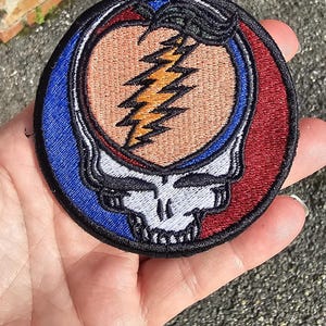 Könnte beinhalten: Ein runder gestickter Aufnäher mit dem legendären Steal Your Face-Logo der Grateful Dead. Das Logo ist ein Schädel mit einem Blitz in der Stirn, umgeben von einem rot-weiß-blauen Design.