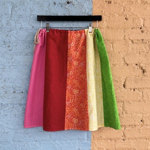 Puede incluir: Una falda de retazos colorida con paneles de tela rosa, rojo, naranja, amarillo y verde. La falda está colgada de una percha negra contra una pared azul y naranja.