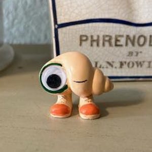 May include: A whimsical figurine of a snail with a large, googly eye and orange shoes. The snail is tan with a spiral shell. The figurine is on a light-colored surface, with a bust and a glass jar with colorful balls in the background.