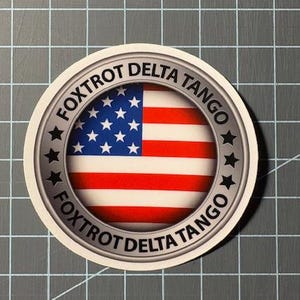 May include: A circular sticker featuring the American flag. The flag is centered within a silver border, with the words "FOXTROT DELTA TANGO" in a circular pattern around the flag. The sticker has a white background.