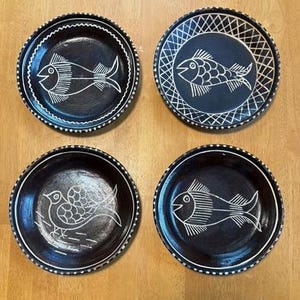 May include: Four black ceramic plates with white etched designs. Two plates feature fish, one a bird, and the last a fish. Each plate has a white border. The plates are arranged on a wooden surface.