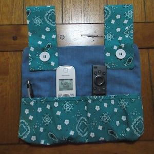 May include: A teal blue fabric organizer with white paisley print and two white buttons. The organizer has three pockets and is hanging on a wooden surface. A white phone with a digital clock displaying "6:45 AM" and a black remote control are in the pockets.