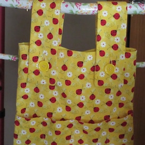 May include: A yellow fabric tote bag with a ladybug and daisy pattern. The bag has two pockets on the front and yellow straps that attach with yellow buttons. The bag is designed to hang on a walker.