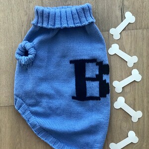 May include: A light blue knitted dog sweater with a black letter "E" on the front. The sweater has a turtleneck and a hole for the leash.