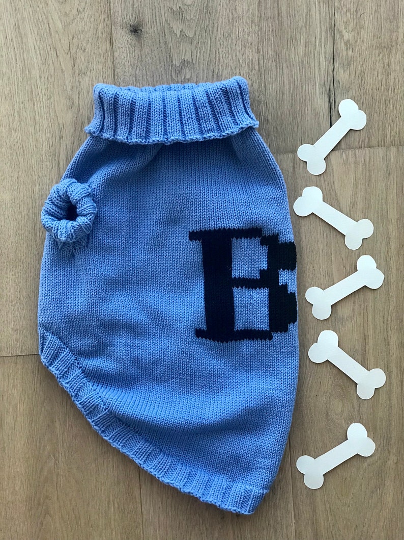May include: A light blue knitted dog sweater with a black letter "E" on the front. The sweater has a turtleneck and a hole for the leash.