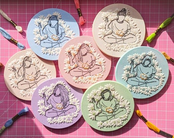 May include: Seven embroidered hoop art pieces featuring a person sitting cross-legged with flowers surrounding them. Each hoop is a different color: white, light blue, pink, light green, lavender, light blue, and beige.