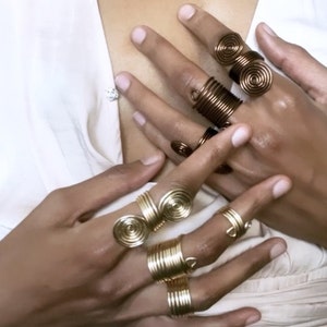 May include: A set of gold and copper wire rings with a spiral design. The rings are worn on multiple fingers.