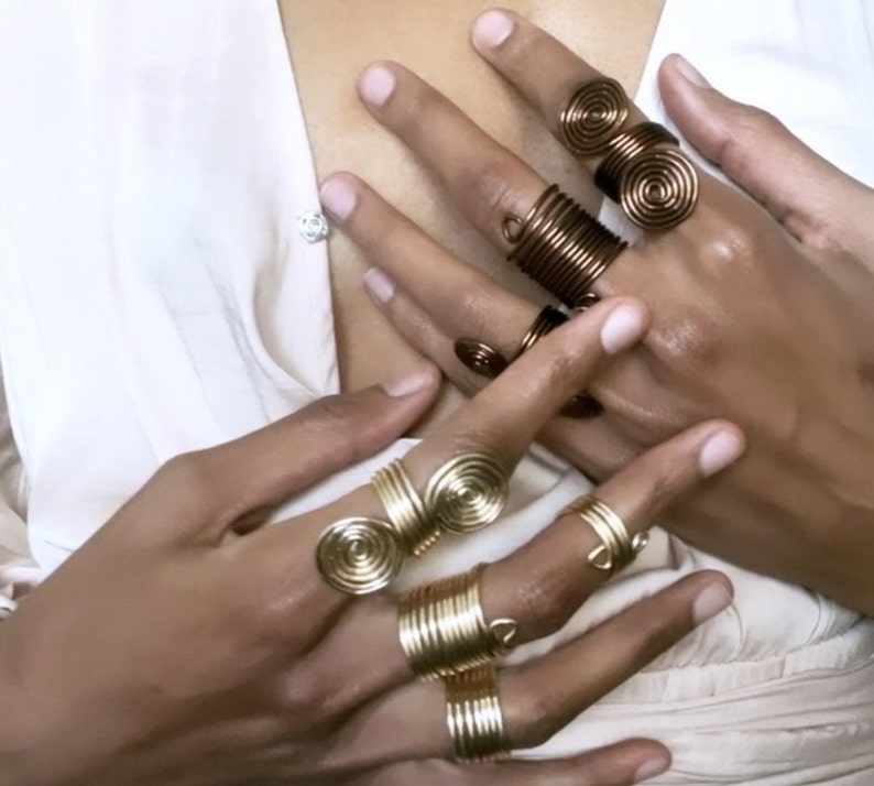 May include: A set of gold and copper wire rings with a spiral design. The rings are worn on multiple fingers.