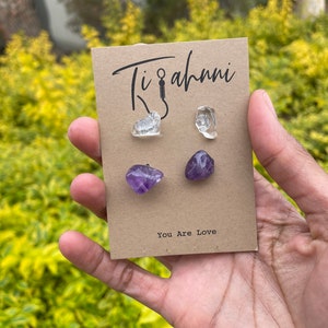 May include: A set of three crystal earrings, two clear quartz and one amethyst. The earrings are displayed on a brown card with the text "Ti'ahni" and "You Are Love".
