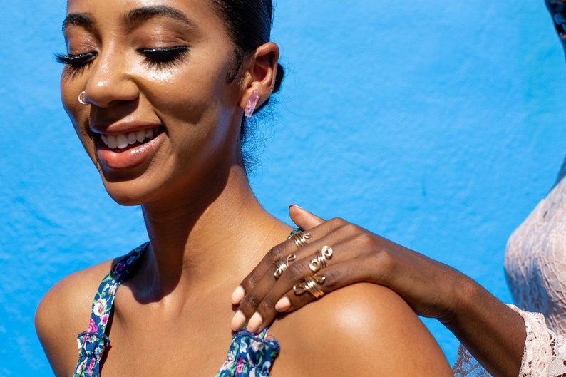 May include: A woman with dark hair and a bright smile wears a floral print top and several gold rings on her fingers. She is standing in front of a bright blue background.