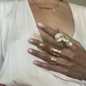 May include: A set of gold-toned rings, including a double spiral ring and several thin, coiled rings. The rings are worn on a person's fingers.