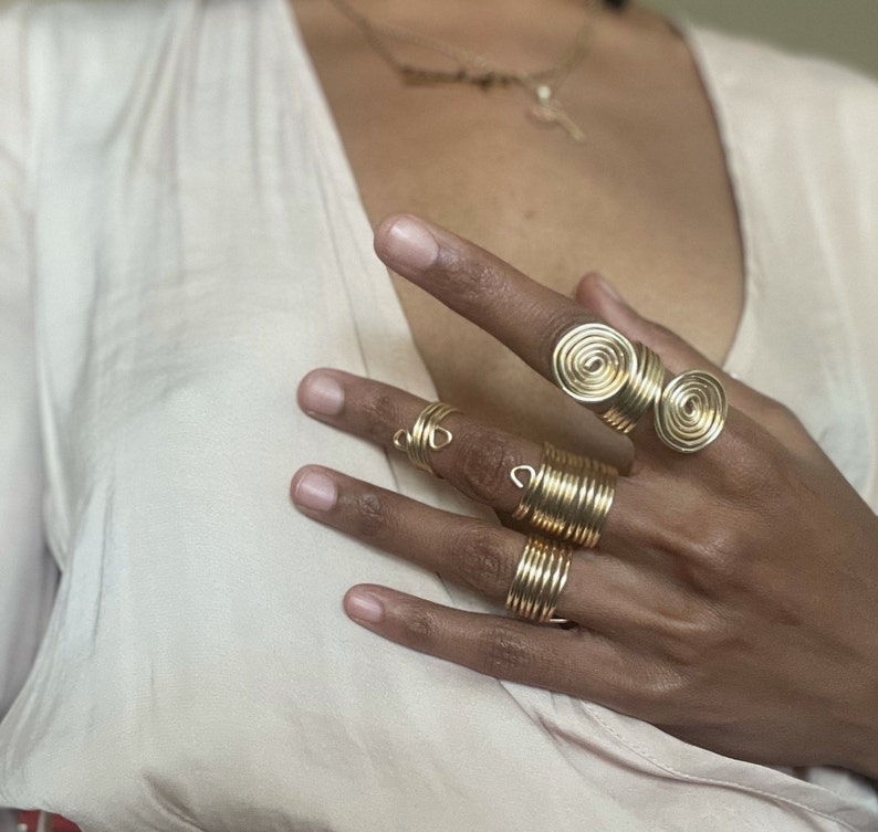 May include: A set of gold-toned rings, including a double spiral ring and several thin, coiled rings. The rings are worn on a person's fingers.