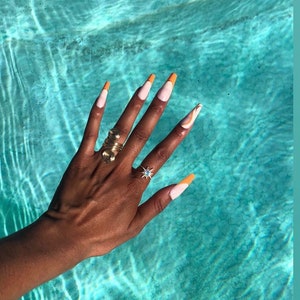 May include: A hand with long, white nails with orange tips. The nails are painted with a white base and an orange tip. The hand is wearing two rings, one gold and one silver. The hand is resting on a blue surface.