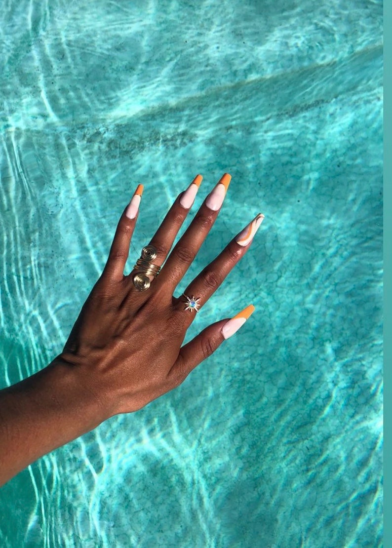 May include: A hand with long, white nails with orange tips. The nails are painted with a white base and an orange tip. The hand is wearing two rings, one gold and one silver. The hand is resting on a blue surface.