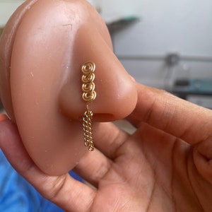 May include: Gold nose ring with a chain dangling from the bottom. The ring is made of multiple small circles.