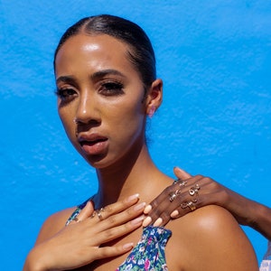 May include: A woman with dark brown hair and skin is wearing a floral print top and gold rings on her fingers. She is looking at the camera with a serious expression. Her hand is resting on her shoulder, and another hand is gently touching her arm.
