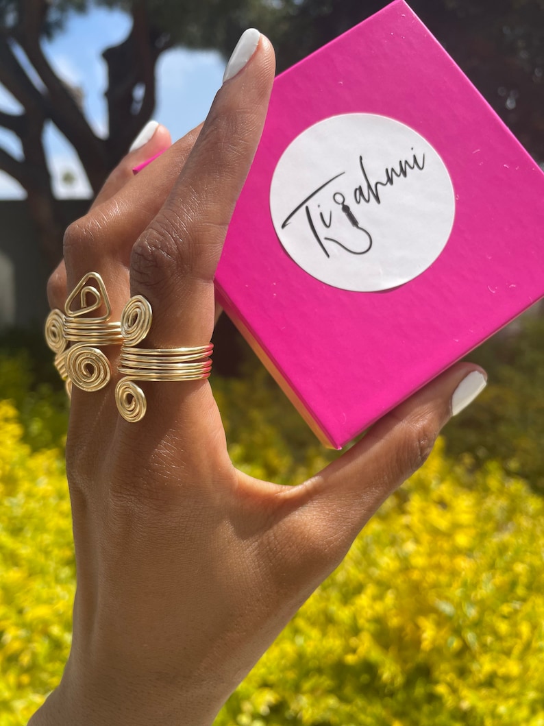 May include: A hand wearing a gold ring set with multiple interconnected circles and a triangle. The hand is holding a pink box with the text "Tizahni" in white.