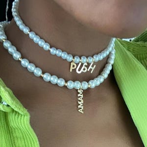 May include: Two-strand pearl necklace with gold accents. The top strand has the word "PUSH" in gold letters, and the bottom strand has a gold pendant that says "AMAM". The necklace is worn against a green shirt.
