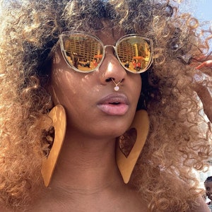 May include: A person with long, curly, light brown hair wearing large, brown heart-shaped earrings and gold-rimmed sunglasses with a clear lens. The person has a nose ring with a small, clear stone.