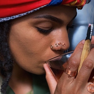 May include: A person wearing a copper wire nose ring and a matching ring on their finger. They are holding a glass of tea and a burning palo santo stick. The person is wearing a colorful headscarf.