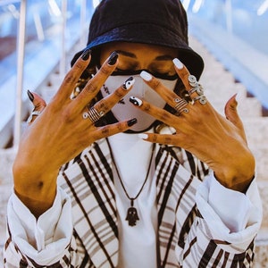 May include: A person wearing a black and white patterned face mask with a black bucket hat and a plaid blazer. The person is wearing several silver rings on their fingers and has black and white nail polish.