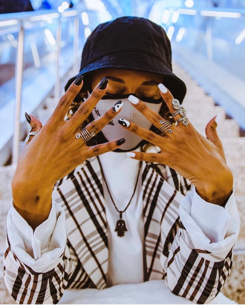 May include: A person wearing a black and white patterned face mask with a black bucket hat and a plaid blazer. The person is wearing several silver rings on their fingers and has black and white nail polish.