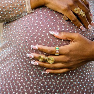 May include: A woman's hands with white nail polish and gold rings on a sparkly, light brown fabric background.