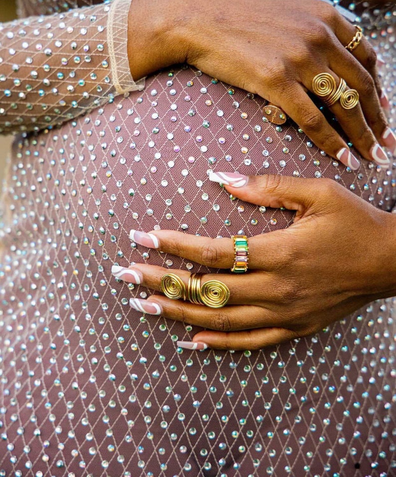May include: A woman's hands with white nail polish and gold rings on a sparkly, light brown fabric background.