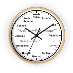 May include: A round wall clock with a light wood frame. The white clock face features black hands, black numerals, and text in a unique font. The clock's design includes the text "Hvshi kanvlliyynanta?"
