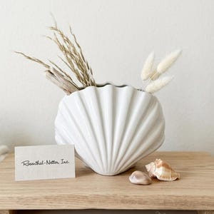 May include: A white ceramic seashell-shaped vase holds dried grasses and fluffy white stems. A small card reads "Rosenthal-Netter, Inc." Two seashells and a small stone sit nearby. The vase is on a wooden tray.