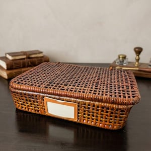 May include: A rectangular, woven rattan basket in shades of brown and orange. The basket features a label holder with a white insert. The basket is on a dark wooden surface, with books and desk accessories in the background.