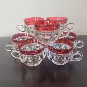 May include: A stack of seven clear glass teacups with ruby red rims and handles. The cups have a decorative pattern around the sides. The teacups are arranged in a pyramid shape, with the bottom row containing three cups.