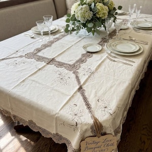 May include: An off-white rectangular banquet cloth featuring intricate brown lace trim and floral embroidery. A wooden tag attached to the corner displays the text Vintage Lace Banquet Cloth (72"x108"). The cloth is set on a table with plates.